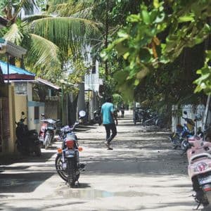 Una persona camina por una calle arenosa bordeada de motocicletas aparcadas, casas y exuberantes árboles tropicales.