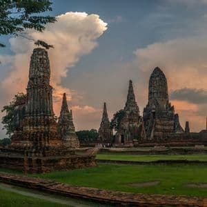 Ancient stone temple ruins with tall spires stand on a grassy field under a cloudy sky at sunset.