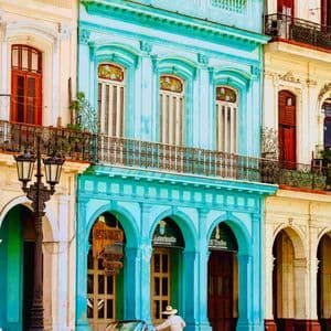 Una persona con sombrero de paja junto a un coche descapotable rosa vintage, aparcado frente a edificios coloridos y ornamentados con balcones.