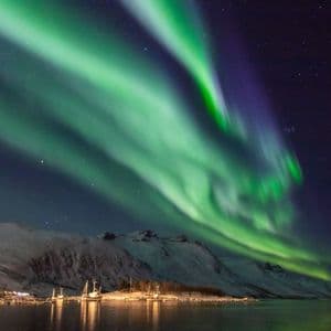 Aurora boreale verde che risplende nel cielo notturno sopra montagne innevate e un porto illuminato con barche.