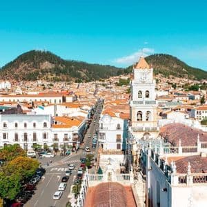 Vista elevada de una ciudad histórica con una torre de reloj blanca, techos de terracota y una calle larga que conduce hacia colinas verdes.