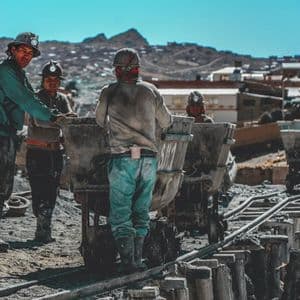 Un grupo de mineros con cascos y lámparas frontales trabaja con vagonetas en una vía férrea al aire libre.