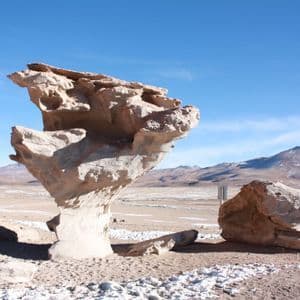 Una gran formación rocosa erosionada por el viento se alza en un desierto arenoso con montañas nevadas a lo lejos bajo un cielo azul claro.