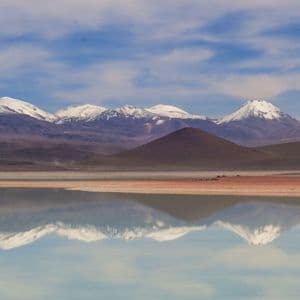 Una cadena de montañas cubiertas de nieve y colinas marrones se reflejan en las tranquilas aguas de un lago bajo un cielo azul con nubes ligeras.