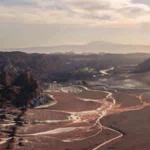 Un vasto valle desértico con oscuras montañas rocosas y sinuosos lechos de ríos secos de color claro bajo un cielo brumoso y luminoso.