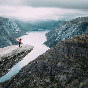 Una persona con una giacca arancione sta a braccia aperte su una cengia rocciosa, affacciata su un fiume che serpeggia in una vasta valle di montagna.