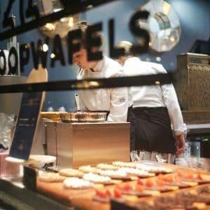 Un chef con uniforme blanco prepara stroopwafels frescos detrás del escaparate de una pastelería lleno de pasteles decorados.