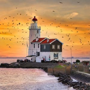 Un faro blanco con techo rojo se alza en una costa rocosa, mientras una bandada de pájaros vuela por el cielo naranja al atardecer.