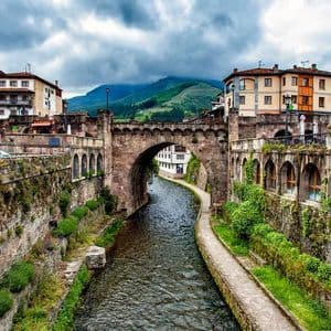 Un puente de arco de piedra cruza un río que fluye a través de un pueblo histórico, con montañas verdes al fondo bajo un cielo nublado.
