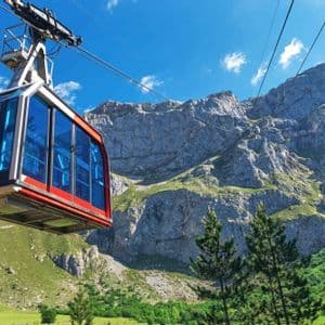 Un teleférico rojo con ventanas azules asciende sobre un valle de montaña verde bajo un cielo azul claro.
