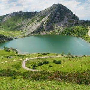 Un lago turquesa se asienta en un valle rodeado de montañas verdes y rocosas, con una carretera sinuosa que bordea su orilla bajo un cielo parcialmente nublado.