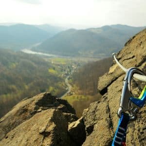 Un mosquetón sujeto a un cable de seguridad de acero en una vía ferrata, en lo alto de un acantilado con vistas a un valle de montaña.