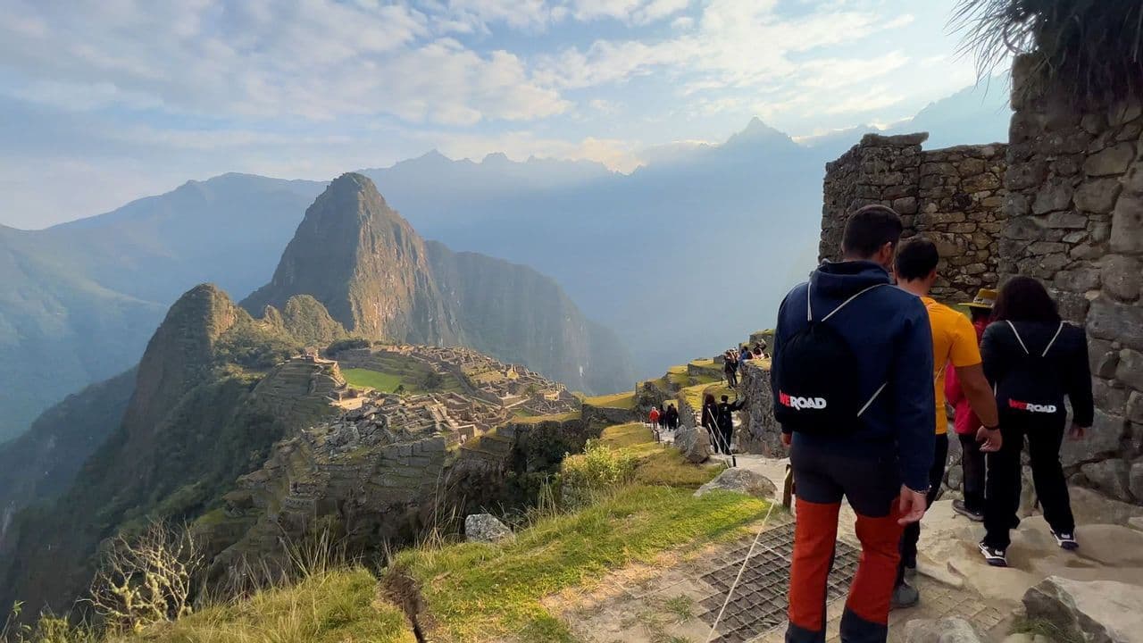 Un viaje en grupo de WeRoad camina por un sendero de piedra, con vistas a ruinas antiguas en la ladera de una montaña bajo un cielo parcialmente nublado.