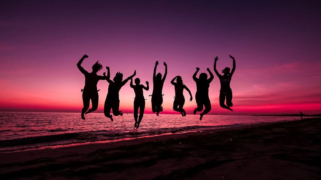 Un voyage de groupe WeRoad avec des silhouettes de personnes sautant sur une plage au coucher de soleil violet et rose.