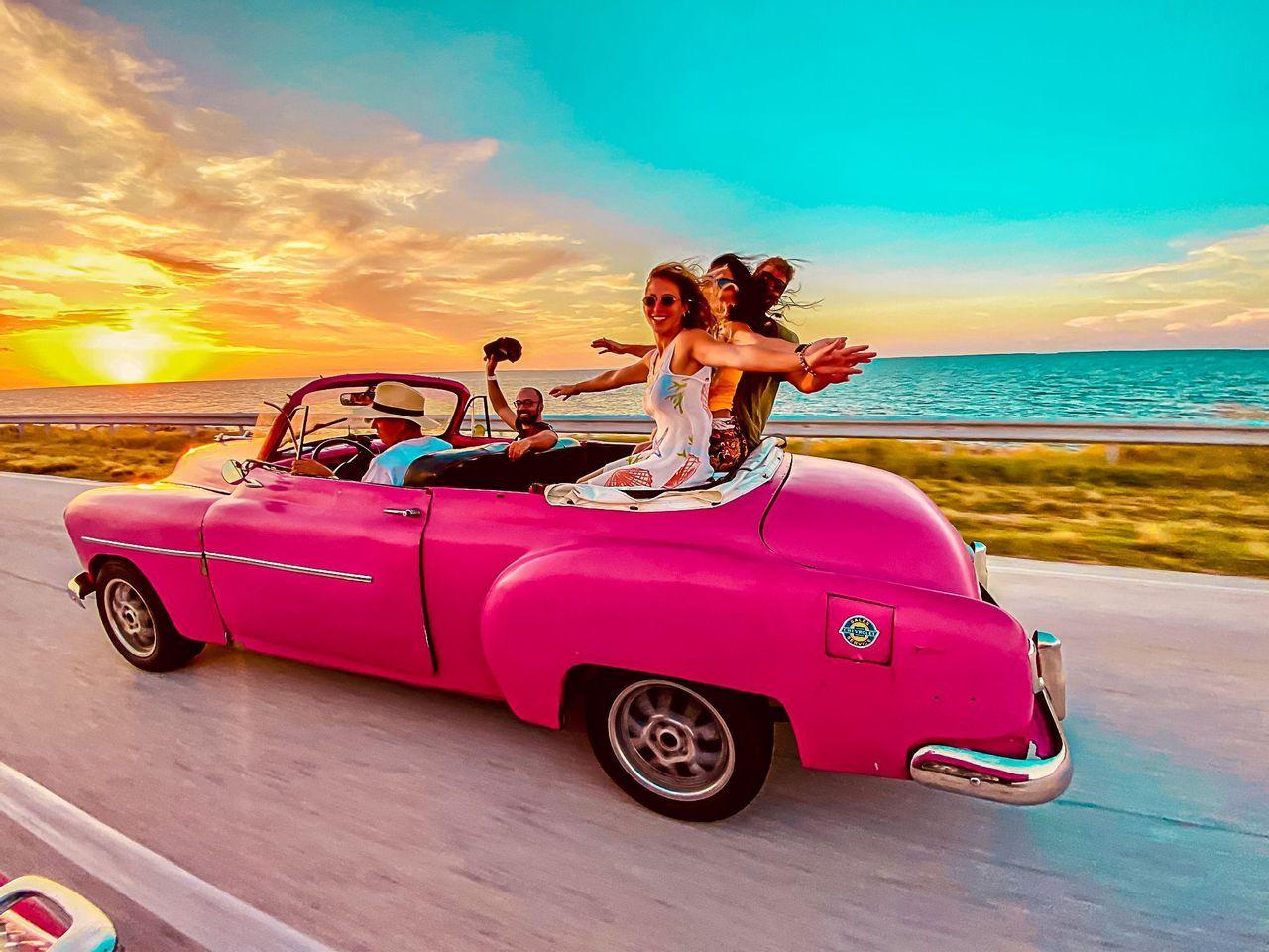 Un grupo de viaje de WeRoad recorre una carretera costera en un clásico descapotable rosa al atardecer, con los brazos extendidos.