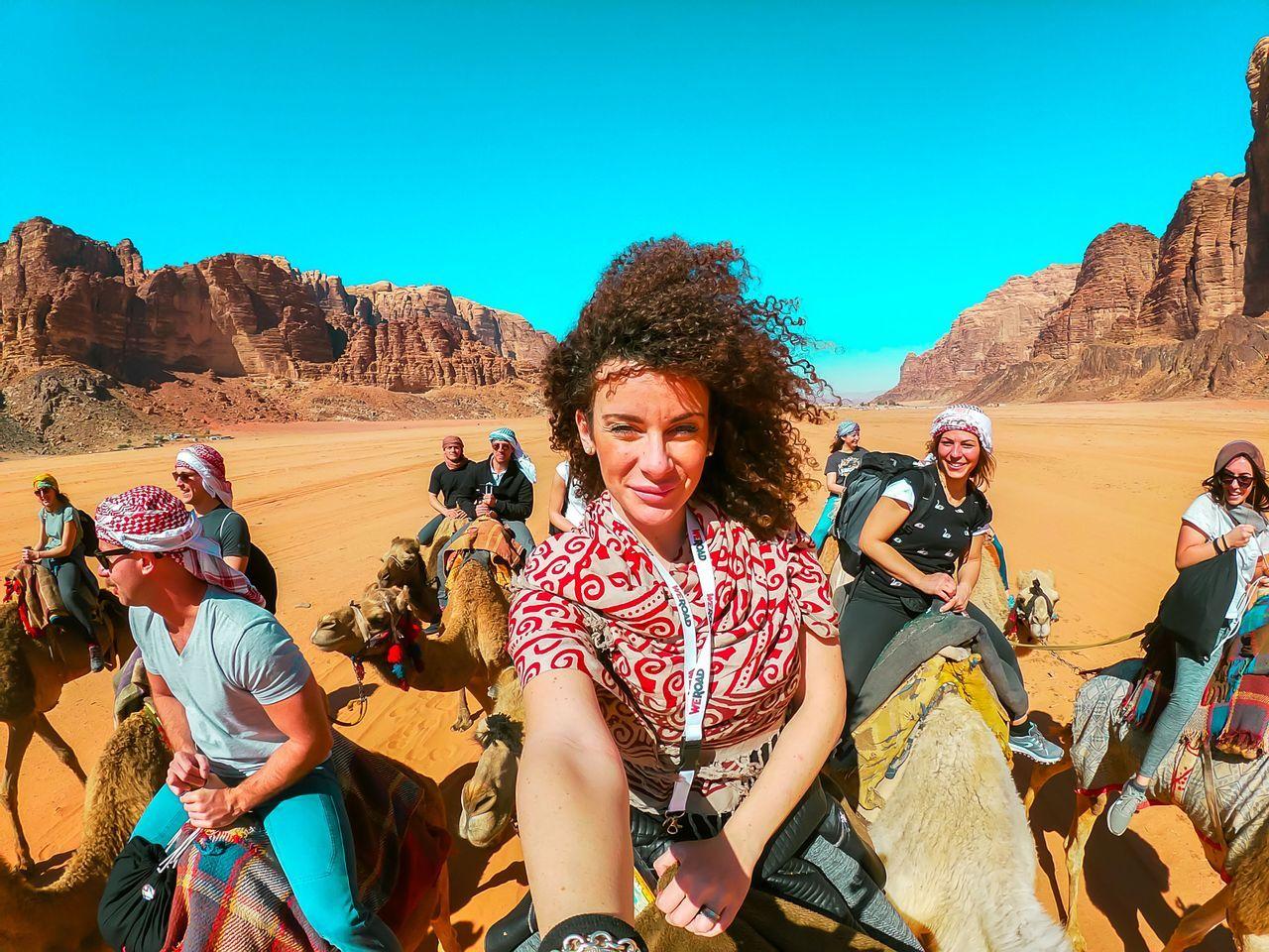 Una mujer se hace una selfie con su grupo de WeRoad mientras monta en camello por un desierto con grandes formaciones rocosas.