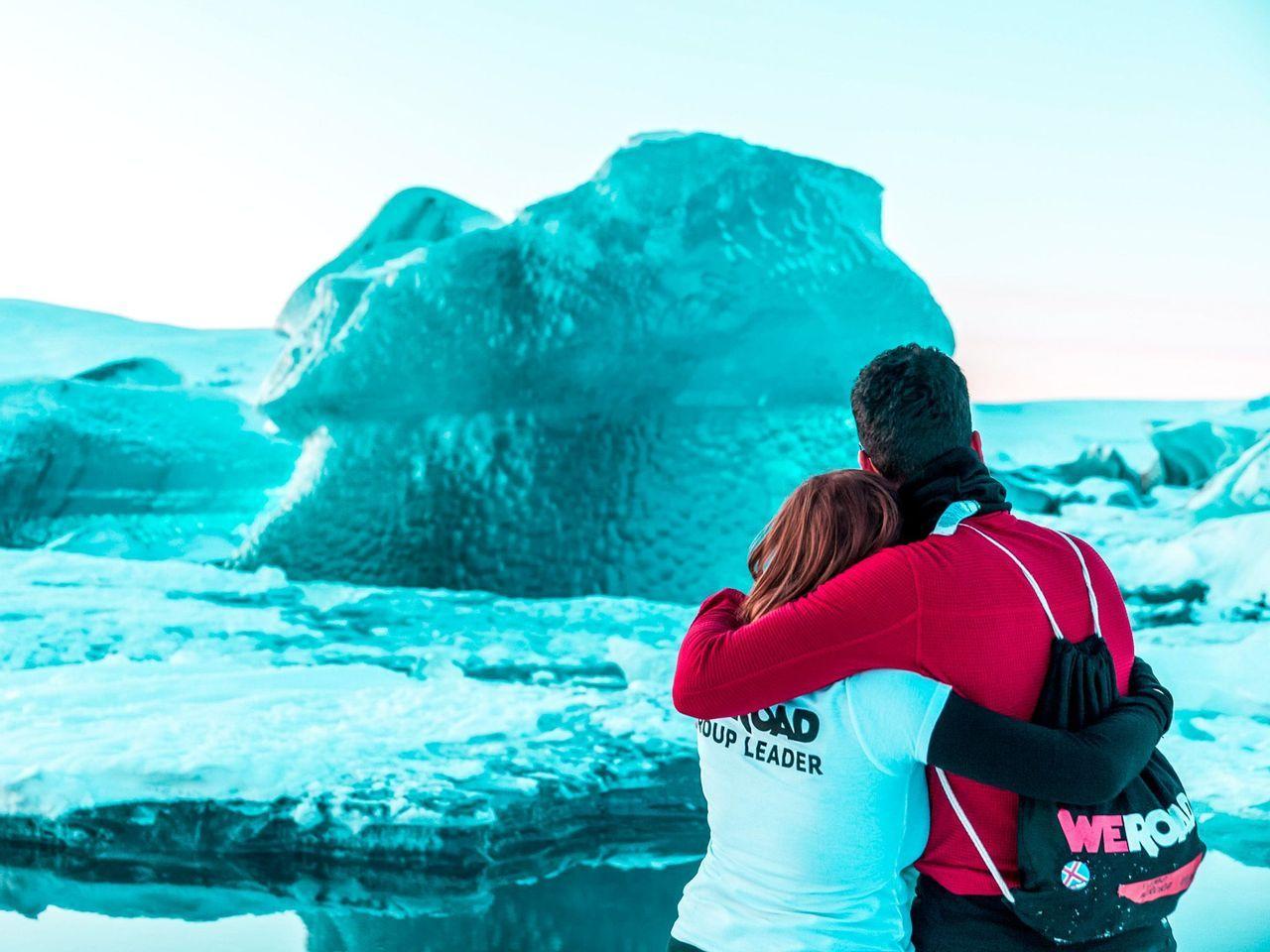 Deux membres d'un groupe WeRoad s'étreignent en contemplant un grand iceberg bleu dans un paysage glacé.