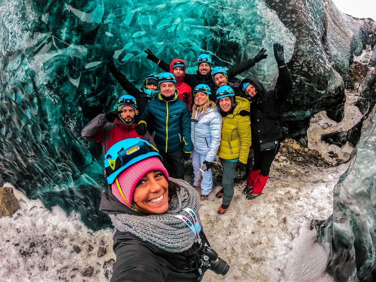 Un viaje en grupo de WeRoad con cascos y chaquetas de invierno se hace una selfie dentro de una cueva de hielo turquesa.