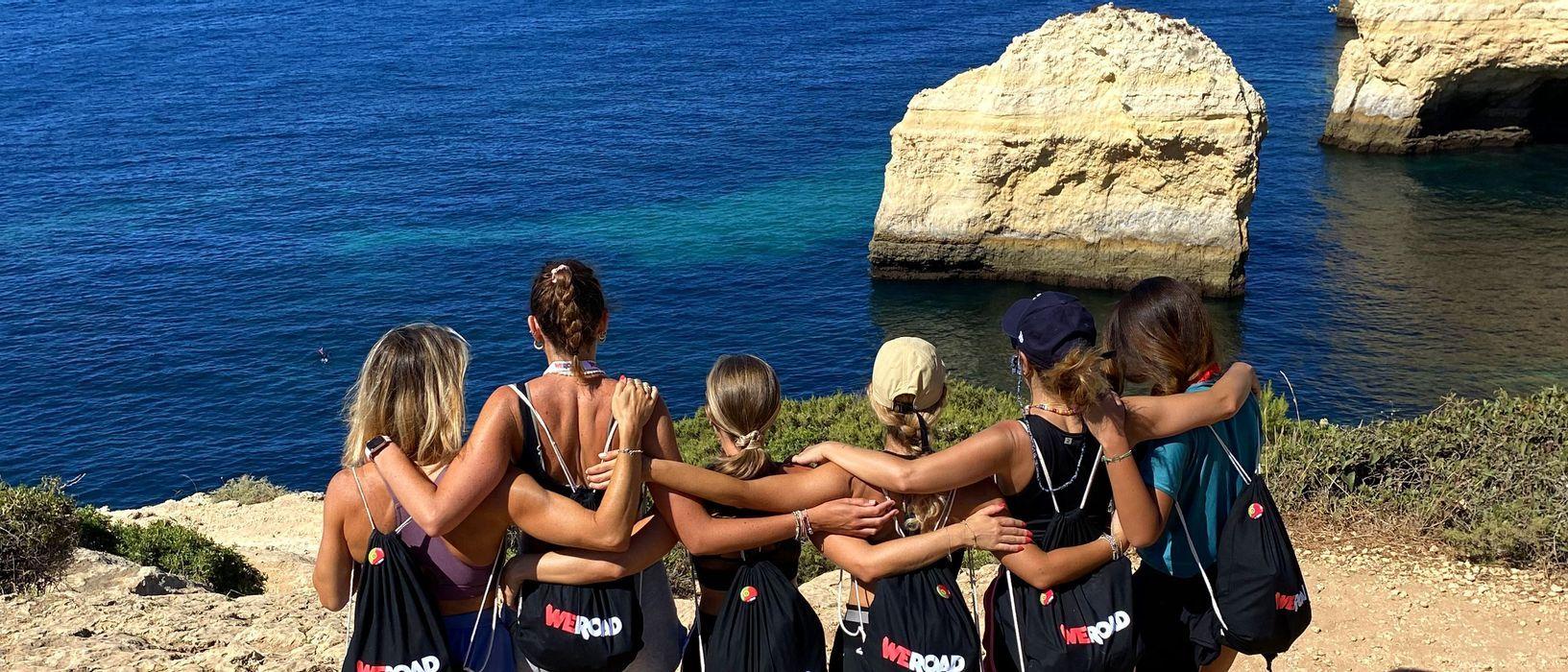 A WeRoad group trip of women stands with arms linked on a cliff, looking out at a blue ocean and coastal rock formations.