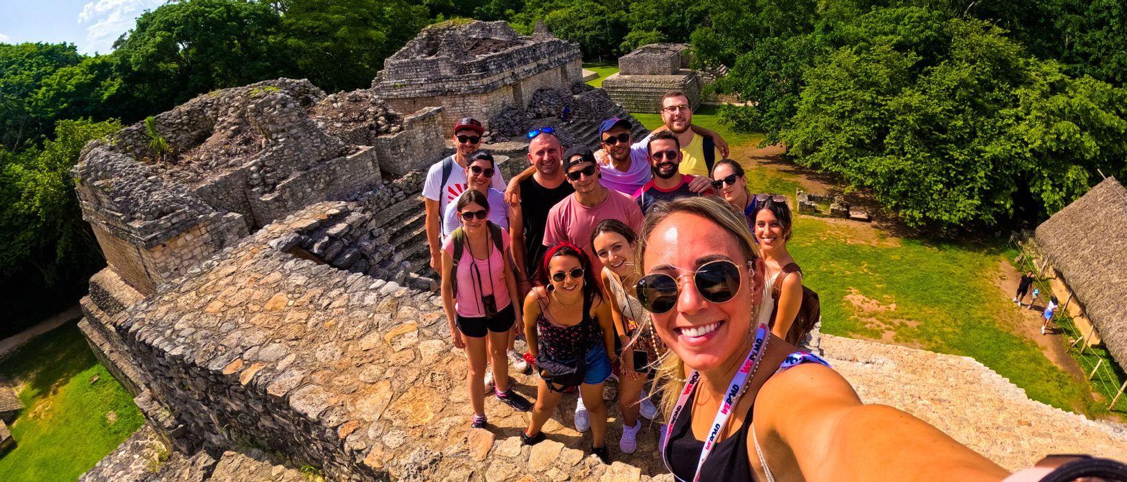 A WeRoad group trip takes a smiling selfie on top of ancient stone ruins with a lush jungle in the background.
