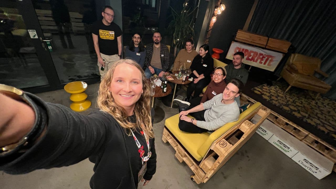 A smiling woman takes a selfie with a WeRoad group posing in a modern lounge with furniture made from wooden pallets.