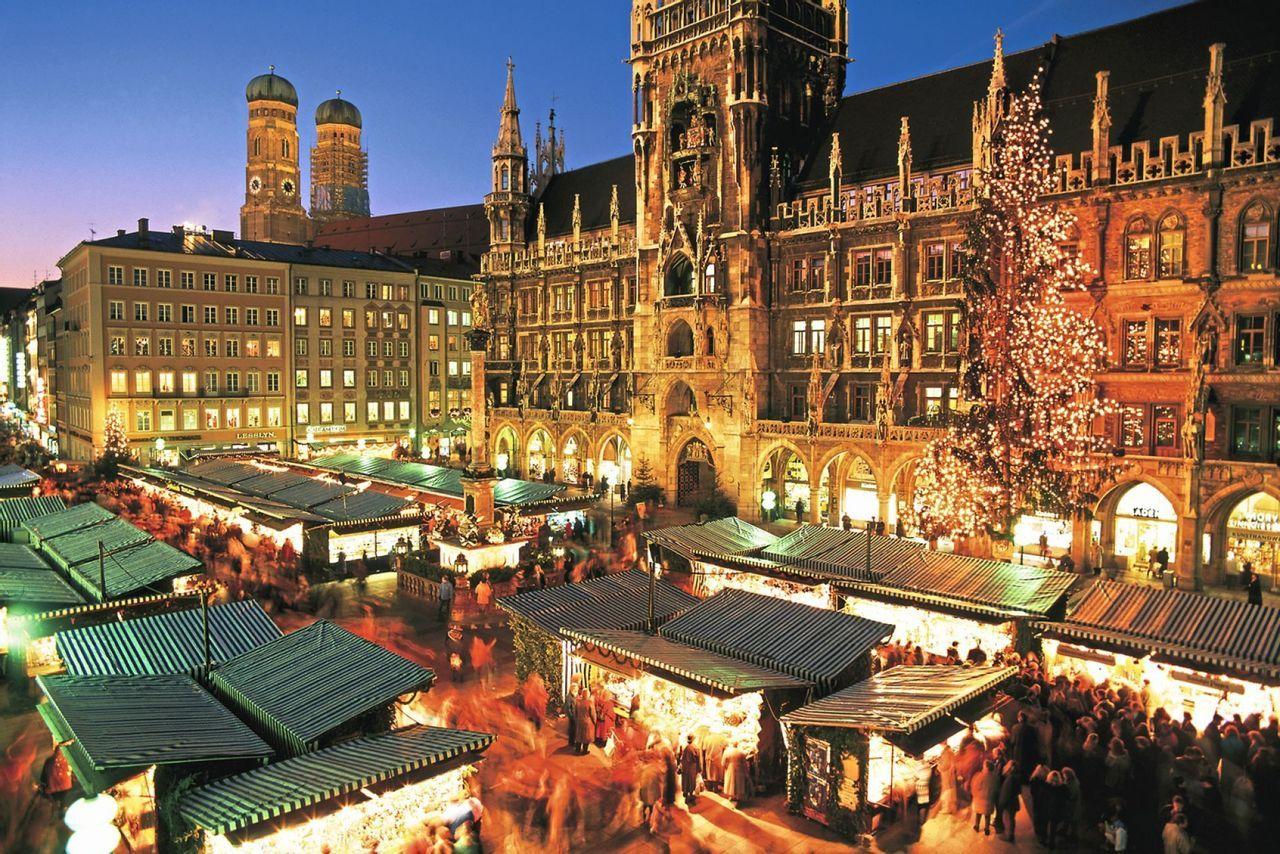 A bustling Christmas market with illuminated stalls and a large Christmas tree fills a historic city square at dusk.