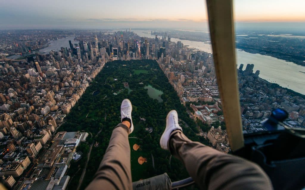 Gambe di una persona in sneakers bianche penzolano da un elicottero, offrendo una vista dall'alto di un vasto paesaggio urbano con un grande parco verde.