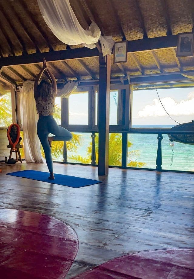 Una donna in posizione dell'albero su un tappetino yoga blu all'interno di uno studio in legno con ampie finestre che si affacciano sull'oceano e sulle palme.