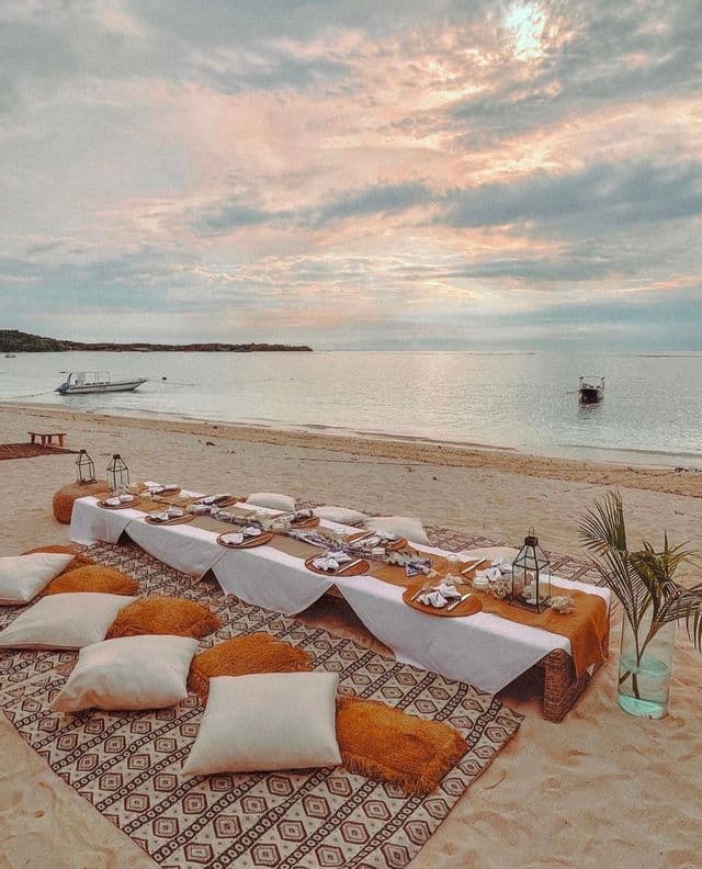 Un tavolino basso con stoviglie e cuscini è allestito per un picnic su una spiaggia sabbiosa in riva all'oceano, sotto un cielo nuvoloso.