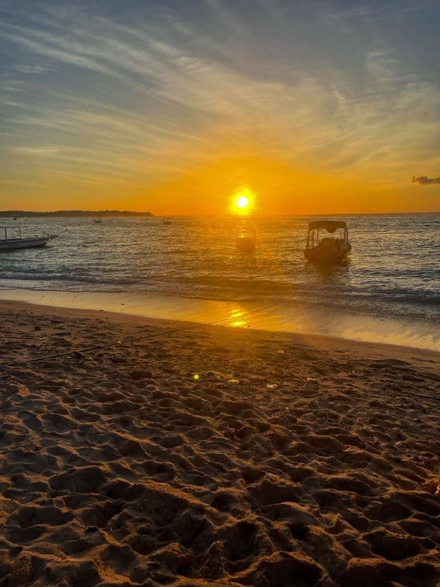 Un tramonto dorato sull'oceano, con barche sull'acqua e una spiaggia sabbiosa in primo piano.