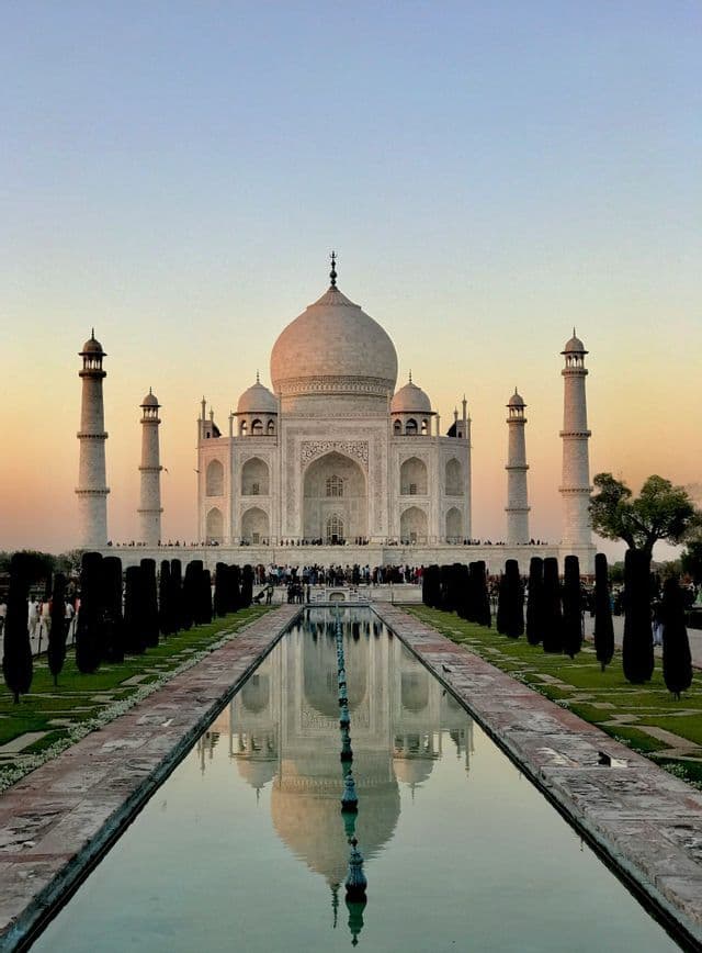 Una vista simmetrica del Taj Mahal che si riflette in una lunga vasca al tramonto, con giardini verde scuro che costeggiano l'acqua.