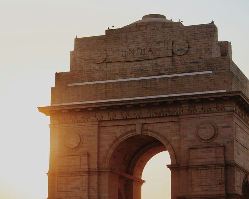 La cima del monumento in pietra India Gate illuminata dalla calda luce del sole al tramonto contro un cielo velato.