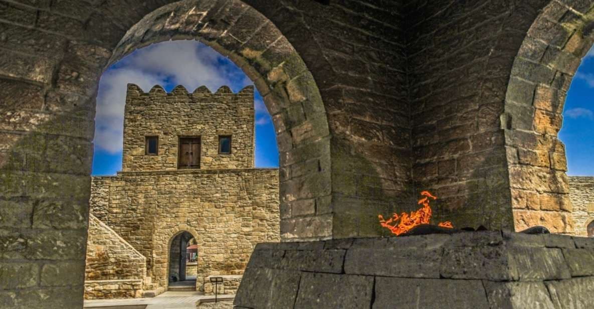 Un fuoco arde su una struttura in pietra sotto un arco, incorniciando la vista di una torre medievale in pietra contro un cielo azzurro.