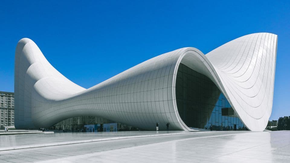 Un grande edificio bianco dal design fluido e curvilineo si trova su un'ampia piazza sotto un cielo azzurro limpido.