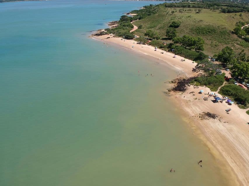 Vista aerea di una spiaggia sabbiosa lungo una costa verde e collinare, con persone che nuotano in acque calme e turchesi.
