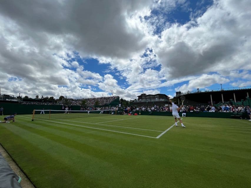 Un tennista in bianco serve durante una partita su un campo in erba, con spettatori sugli spalti sotto un cielo parzialmente nuvoloso.