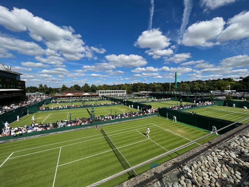 Vista dall'alto di numerosi campi da tennis in erba con partite in corso e spettatori, in una giornata soleggiata e nuvolosa.