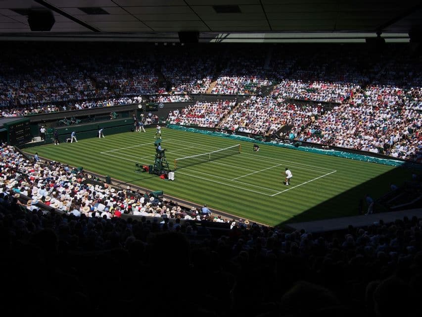Una vista dall'alto di una partita di tennis su un campo in erba illuminato dal sole, con spettatori che riempiono le tribune di un grande stadio.