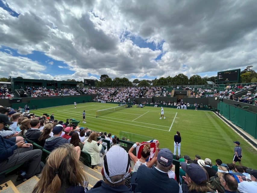 Una folla di spettatori osserva una partita di tennis su un campo in erba dalle tribune sotto un cielo nuvoloso.