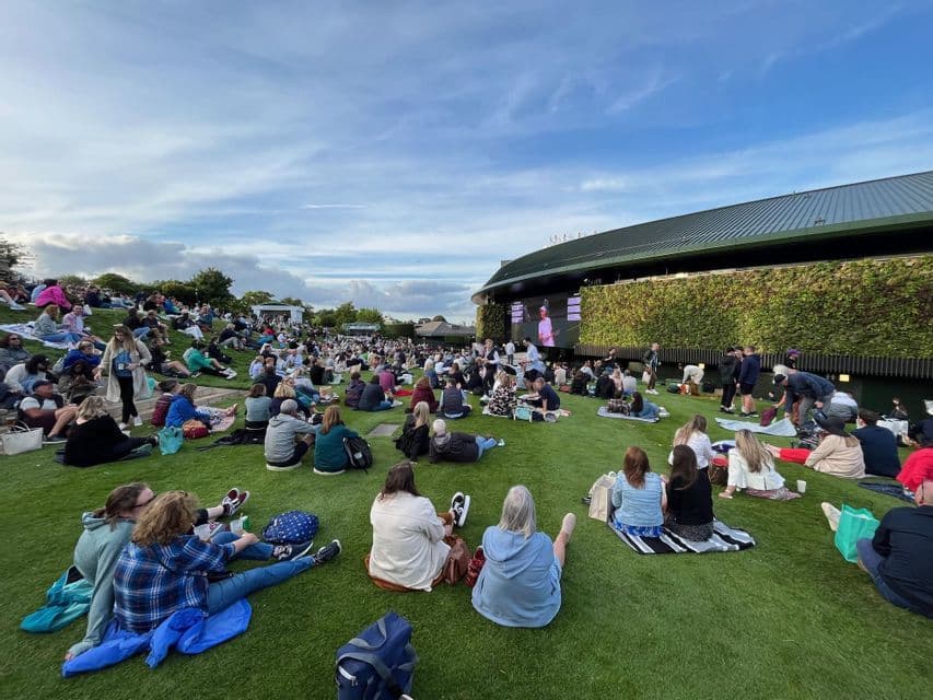 Una grande folla siede su un pendio erboso assistendo a un evento su un grande schermo all'aperto, fissato a un edificio con una parete verde.