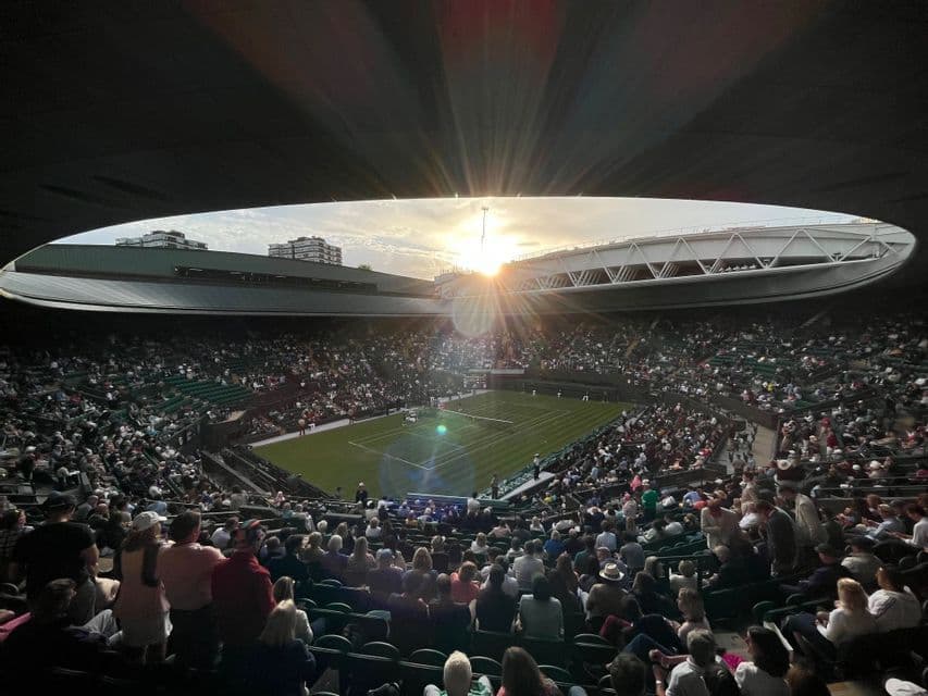 Spettatori guardano una partita di tennis su un campo in erba dalle tribune di un grande stadio con un tetto parzialmente aperto al tramonto.