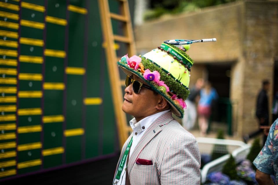 Un uomo in blazer a righe e occhiali da sole indossa un cappello elaborato a tema Wimbledon mentre guarda un tabellone segnapunti.