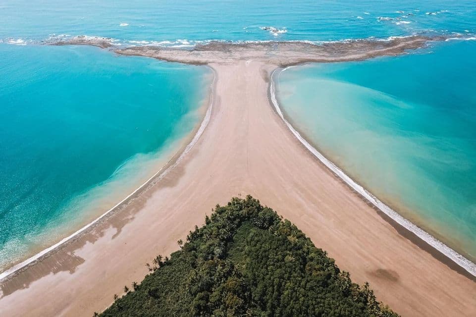 An aerial view of a whale tail-shaped beach formation with turquoise water on both sides and a green forest at its base.