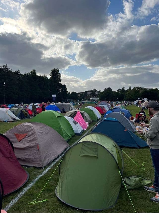 Decine di tende colorate piantate in file su un campo erboso in un campeggio sotto un cielo parzialmente nuvoloso.