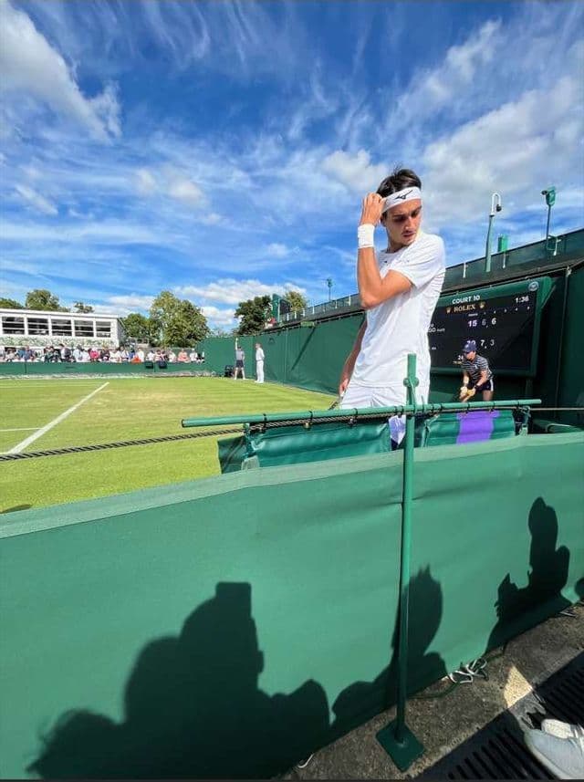 Un tennista in tenuta sportiva bianca si trova su un campo in erba, guardando verso la telecamera mentre si sistema la fascia per capelli.