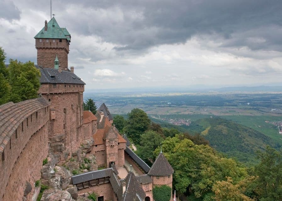 Un castillo de piedra roja con torres y muros fortificados domina un vasto valle verde desde una colina boscosa bajo un cielo nublado.