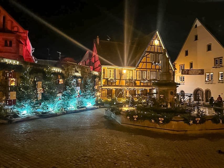 Una plaza de pueblo adoquinada de noche, con edificios de entramado de madera y una fuente decorada con árboles de Navidad y luces festivas.
