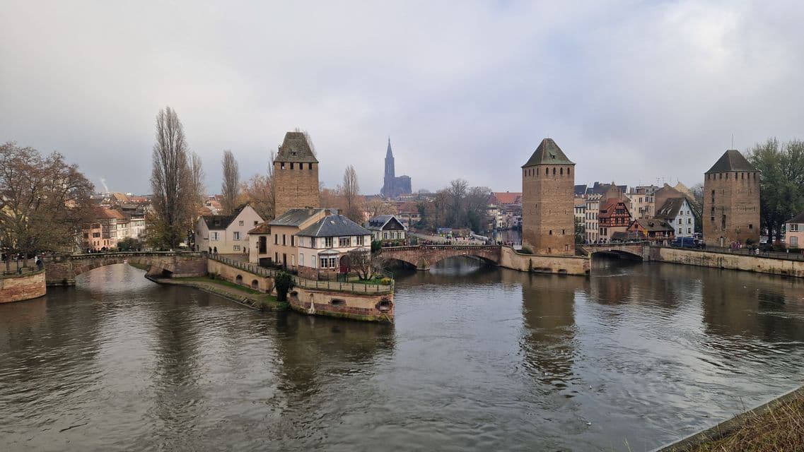 Torres de piedra y puentes arqueados cruzan un amplio río que atraviesa una ciudad histórica, con una catedral visible al fondo.