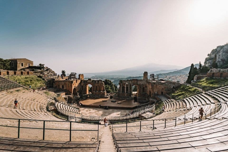 A wide view of an ancient stone amphitheater with tiered seating overlooking the sea and a mountain in the distance.