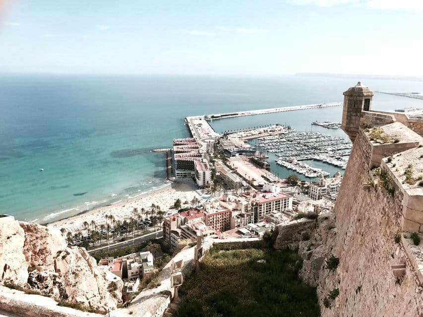Veduta dall'alto da una fortezza in pietra che domina una città costiera, un porto turistico animato con imbarcazioni e una spiaggia di sabbia.
